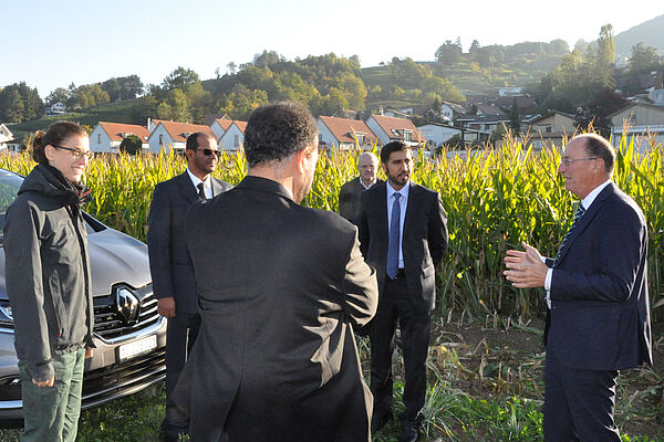 A group of people from Switzerland and Abu Dhabi stand on a field in the morning sun. The group looks towards Urs Niggli on the right side, while he is explaining something. The group is standing on grass, behind them is open soil followed by a maize field. In the background are some village houses and a hill.