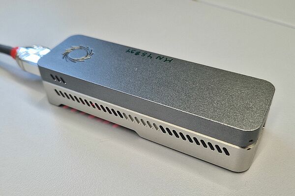 A small, elongated, silver device can be seen. It is lying on a table, has a circular logo and ventilation slots on the side.