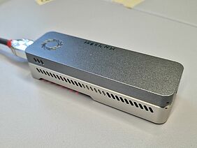 A small, elongated, silver device can be seen. It is lying on a table, has a circular logo and ventilation slots on the side.