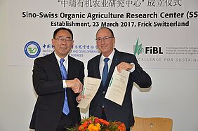 Two men shake hands and hold up a signed contract.