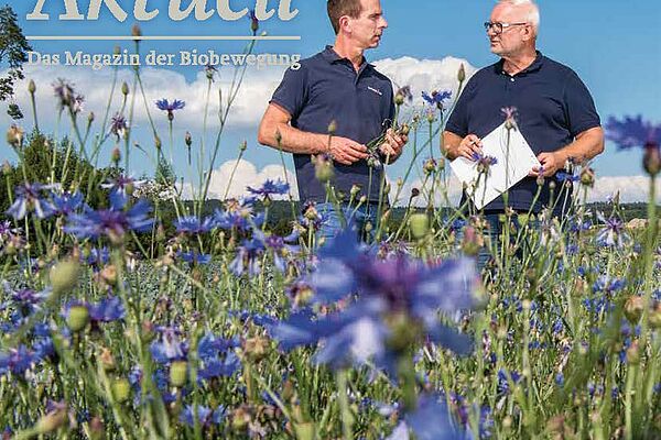 Titelseite Bioaktuell, zwei Männer im Gespräch auf einem Feld, im Vordergrund blaue Blumen.