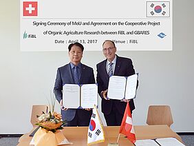 2 men stand at a table and present the signed agreements. THe table is decorated with a Swiss and a South Korean flag as well as some flowers.