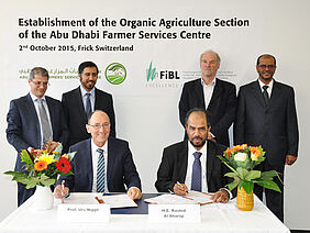 Professeur Docteur Urs Niggli et Son Excellence Rashid Al Shariqi sont assis à une table, qui est décorée avec des fleurs. Les deux hommes tiennent des stylos dans leurs mains, prêts à signer le contrat. Les membres du FiBL et de l’Abu Dhabi Farmers’ Services Centre se tiennent derrière eux. Le mur est couvert par une affiche intitulée „Establishment of the Organic Agriculture Section of the Abu Dhabi Farmers’ Services Centre, 2 October 2015, Frick Switzerland ».