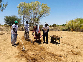 Vier dunkelhäutige Menschen stehen auf einem gelben, trockenen Feld in Afrika. Im Hintergrund ist ein Baum und weiter hinten Gebüsch zu sehen. Die Menschen bearbeiten den Boden mit Hacken und verteilen Mulch darauf.