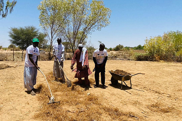 Vier dunkelhäutige Menschen stehen auf einem gelben, trockenen Feld in Afrika. Im Hintergrund ist ein Baum und weiter hinten Gebüsch zu sehen. Die Menschen bearbeiten den Boden mit Hacken und verteilen Mulch darauf.