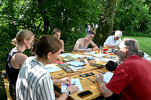 Personen sitzen an einem Tisch mit Malutensilien und malen.