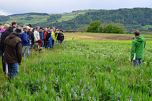 Eine grosse Gruppe von Menschen steht in einem Feld und hört dem Sprecher zu