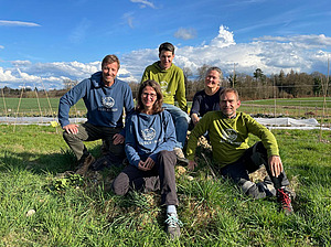 Three men and two women are posing together in front of a vegetable field. They are all wearing jumpers bearing the farm's logo. The name 'Glück-Hof' is written in white against a blue or green background.