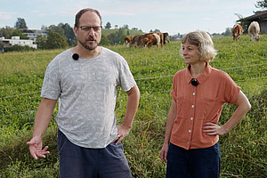 Ein Mann und eine Frau mit Kühen im Hintergrund.