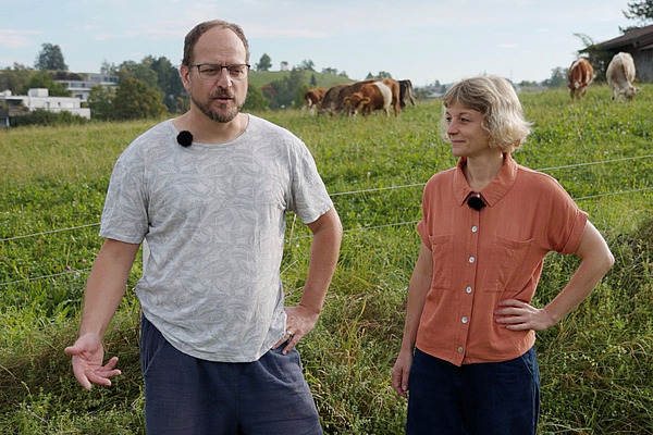 Ein Mann und eine Frau mit Kühen im Hintergrund.