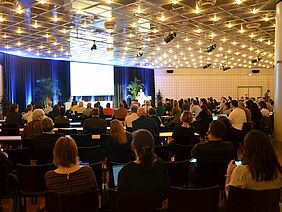A lighted room with many full rows of tables, a speaker standing in front and a presentation on the wall to his left