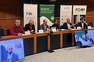 Six people at a podium with two people on screens.