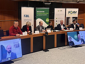 Six people at a podium with two people on screens.