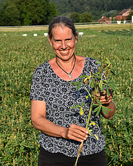 Eine Frau um die 40 oder 50 lächelt sehr freundlich in die Kamera. Sie steht auf einem Lupinenfeld und hält eine der krautigen Pflanzen in der Hand. Es ist sehr sonnig.