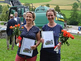 Deux femmes avec des bouquets de fleurs et des certificats devant un excavateur.