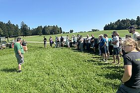 Eine Gruppe von Menschen steht rechts im Bild, link ein einzelner Herr. Dazwischen ein grün bewachsenes Feld.