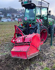 Ein Traktor mit einer Maschine zum Spaten fährt auf einem Feld. Das Wetter ist winterlich grau. Die Maschine ist grell rot.