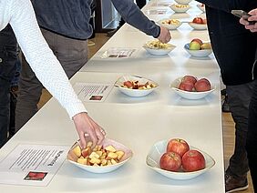 Une table avec des pommes, entières et coupées, dans des coupes. Des personnes se tiennent autour de la table et saisissent parfois les pommes.