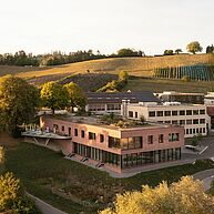 ZU sehen ist ein zweistöckiges, modernes Gebäude und ein kleiner Gebäudekomplex im Hintergrund. Rundherum ist idyllische, grüne Landschaft mit Feldern und Bäumen. Das Licht ist sehr weich und es herrscht Abendstimmung.