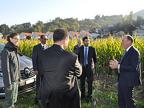 A group of people from Switzerland and Abu Dhabi stand on a field in the morning sun. The group looks towards Urs Niggli on the right side, while he is explaining something. The group is standing on grass, behind them is open soil followed by a maize field. In the background are some village houses and a hill.