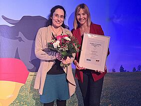 Zwei Frauen mit einem Blumenstrauss und einer gerahmten Auszeichnung in den Händen lächeln in die Kamera und stehen vor einem Poster des Bundesverbands Deutscher Milchviehhalter.