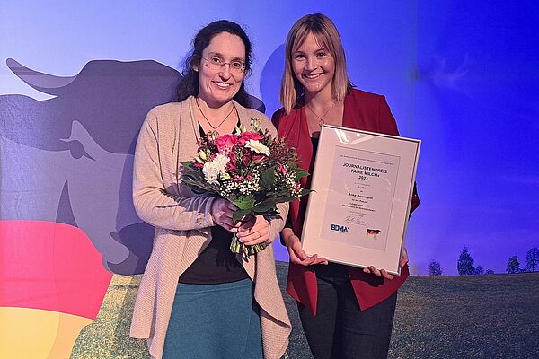 Zwei Frauen mit einem Blumenstrauss und einer gerahmten Auszeichnung in den Händen lächeln in die Kamera und stehen vor einem Poster des Bundesverbands Deutscher Milchviehhalter.