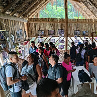 In an open, wooden hut a group of south americans is looking ath pictures on the walls. Through the open part of the hut wall the rainforest is visible. People have cameras and are talking to each other.