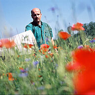 Ein Mann mit einem Kescher steht in einem Feld mit knallroten Mohnblumen. Er hat wenig Haare und ist im mittleren Alter. Der Vordergrund ist unscharf, was dem Bild viel Tiefe gibt.