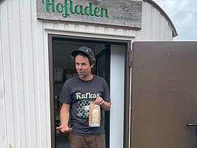 Un homme devant une caravane de chantier avec un panneau "Hofladen" (magasin à la ferme).