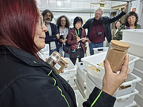 A woman holding chicken feed in a container, people in the background.
