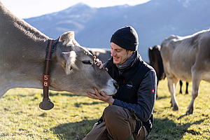 Mann kniet auf einer Wiese und streichekt eine Kuh am Kopf.