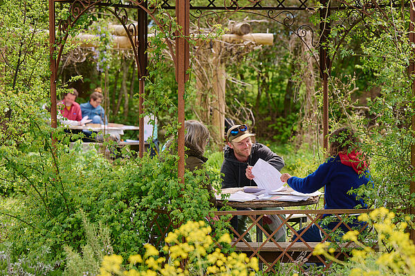 Menschen sitzen an Holztischen in einem Naturgarten.