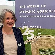 Eine freundlich lächelnde Frau mit schwarzem Blazer und kinnlangen, angegrauten Haaren steht vor einem Banner "25 Years the world of organic agriculture".