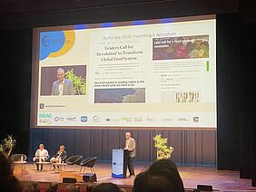 A speaker at the podium in front of a presentation