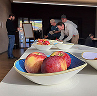 Im Vordergrund ist eine Schale mit sehr schönen Äpfeln auf einem Tisch. Im Hintergrund sieht man Menschen über den Tisch gebeugt, bei einer Degustation in einem Innenraum.