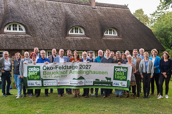 Gruppe Menschen vor einem Haus mit Reddach, die ein Werbebanner für die Öko-Feldtage 2027 halten.