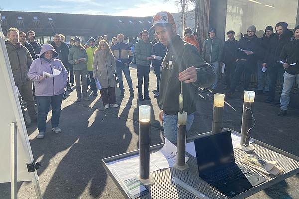 Teilnehmende der Tagung verfolgen im Freien eine Demonstration, bei der verschiedene Boden- oder Flüssigkeitsproben in transparenten Zylindern gezeigt und erläutert werden.