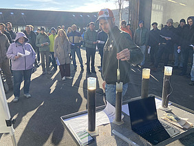 Gruppe von Menschen im Freien, die einem Mann zuschauen, der etwas in zylindrischen Gefässen zeigt. 