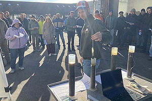 Gruppe von Menschen im Freien, die einem Mann zuschauen, der etwas in zylindrischen Gefässen zeigt. 