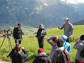  A group of young people listen to three speakers on an alpine meadow.