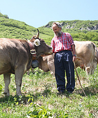Ein urig aussehnder Mann mit einem Stock in der Hand steht auf einer idyllischen Alpweide und krault eine Kuh am Kinn. Die Kuh streckt sich genüsslich zur Hand hin.