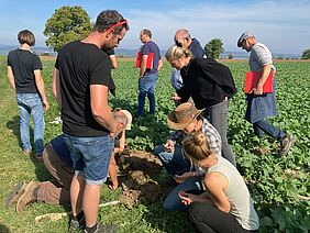 Frauen und Männder schauen sich Bodenproben auf dem Feld an.