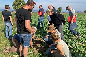 Frauen und Männder schauen sich Bodenproben auf dem Feld an.