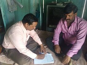 Two men sitting at the floor, one with some papers.