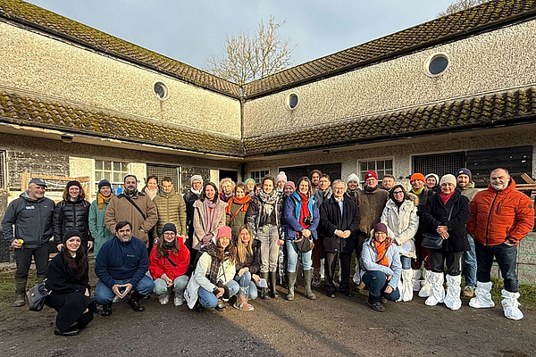 Gruppenfoto von 35 Personen vor einem Gebäude mit Moosdach. Sie sind warm gekleidet, einige tragen Schuhüberzüge. Die Stimmung wirkt freundlich und gemeinschaftlich.