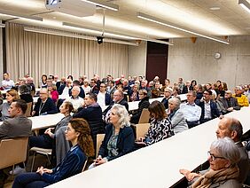 Ein Saal mit mehreren Reihen von Tischen, an welchen viele Leute sitzen.