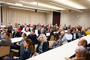 Ein Saal mit mehreren Reihen von Tischen, an welchen viele Leute sitzen.