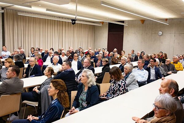 Ein Saal mit mehreren Reihen von Tischen, an welchen viele Leute sitzen.