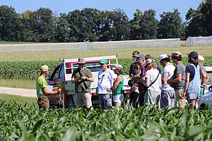 Several people are standing on a dirt track between a maize field and a red vehicle.