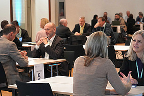 The picture shows tables, each with two to four people sitting at them and talking to one another. The situation appears to be formal.